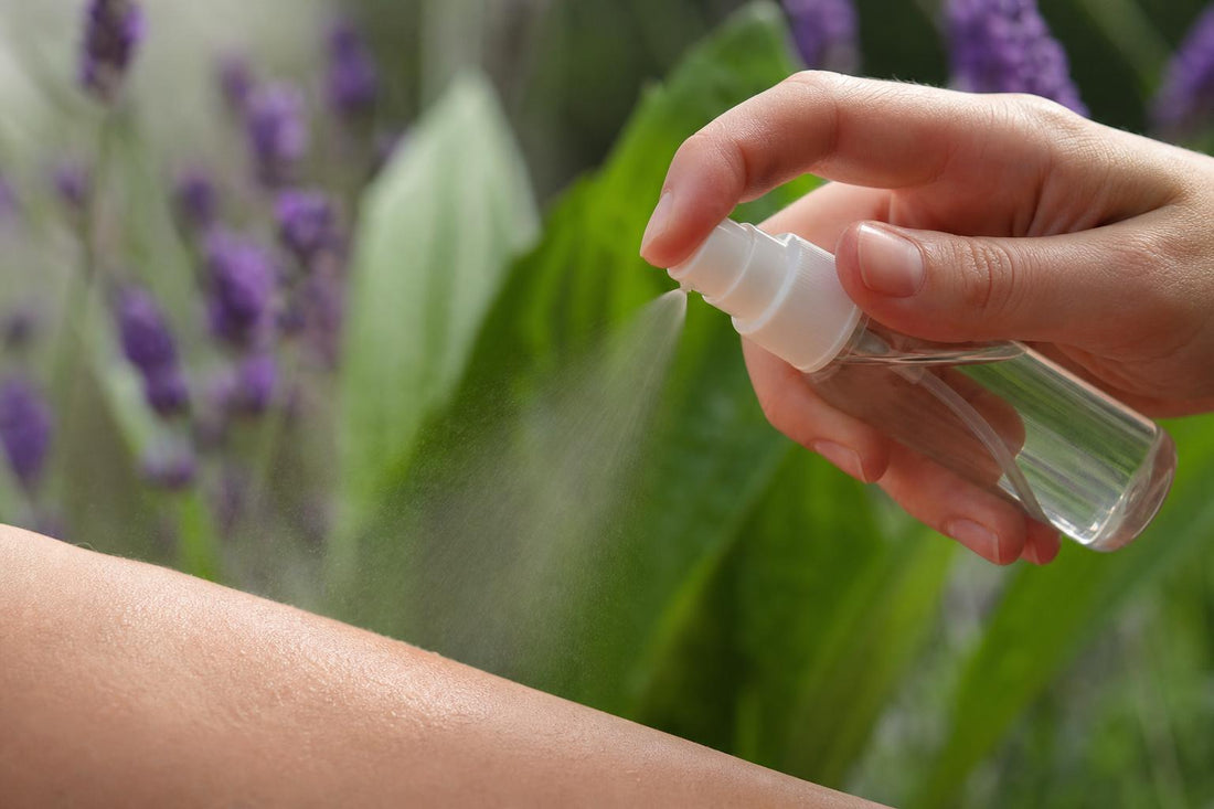 Main appliquant un spray anti-moustiques en extérieur, avec des feuilles de plantain et des fleurs de lavande en arrière-plan flou