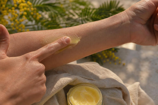 Application d’un baume artisanal sur la peau après le soleil, texture crémeuse visible, ambiance naturelle avec lumière douce et feuillage en arrière-plan.